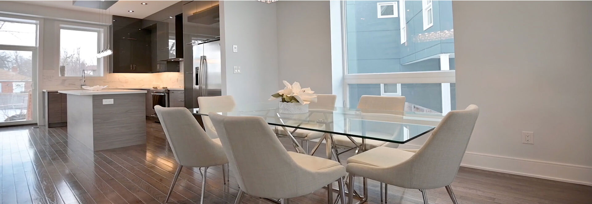 Modern dining area with glass table surrounded by eight beige upholstered chairs and a kitchen with dark cabinets and stainless steel appliances.