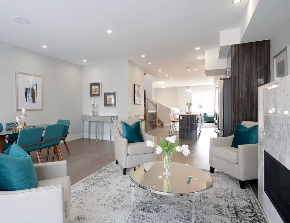 Modern open living room with beige armchairs, teal cushions, round glass coffee table, and a dining area with teal chairs.