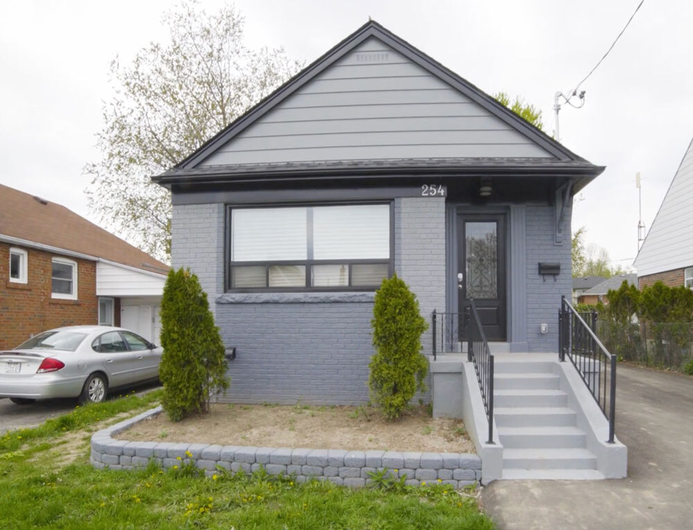 Small gray brick house with front steps, two evergreen shrubs, and a driveway with a silver car parked.