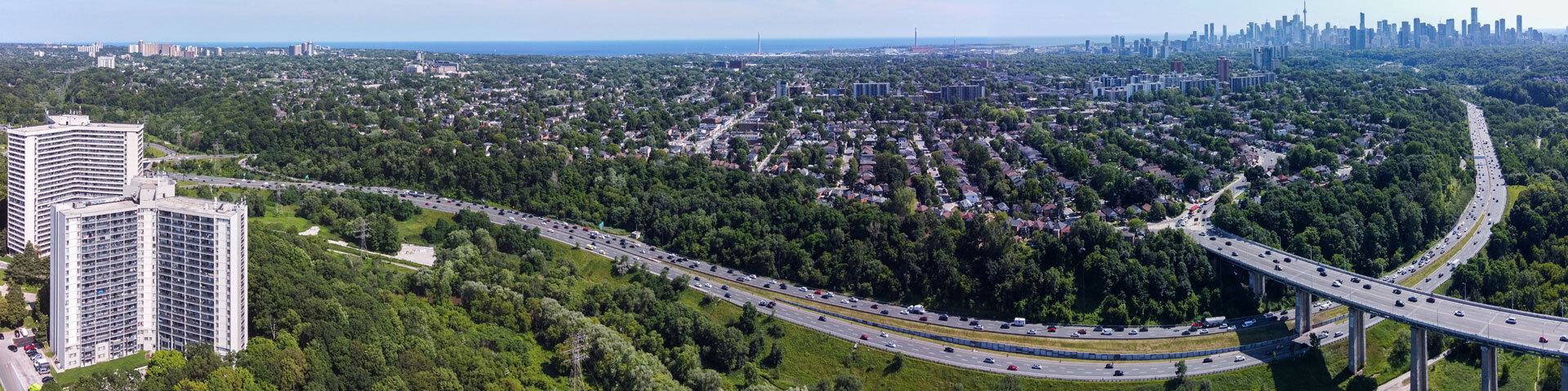 Aerial view of a highway surrounded by dense green trees, residential areas, and a distant city skyline under a clear blue sky.