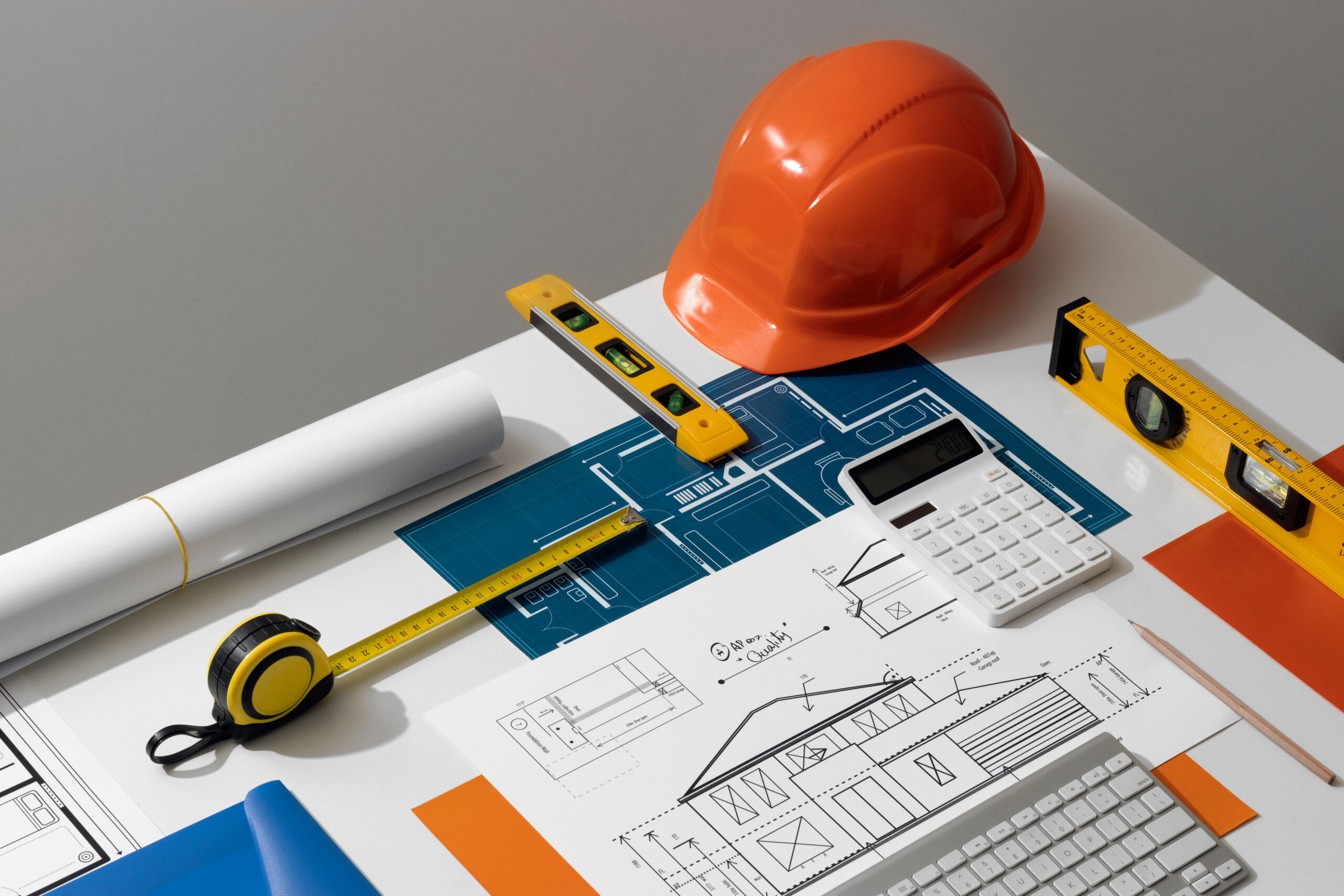 Architectural blueprints and sketches on a table with an orange hard hat, measuring tape, spirit levels, calculator, pencil, and keyboard.