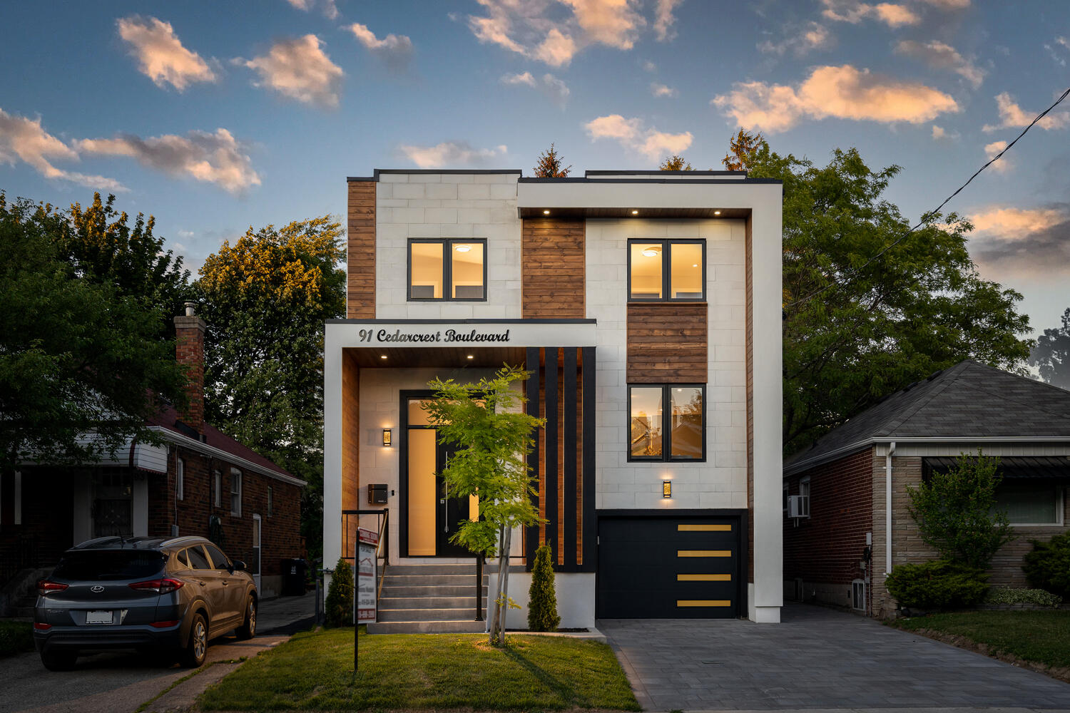 Modern two-story house with wood and white panel exterior, illuminated entrance, and black garage door at dusk.