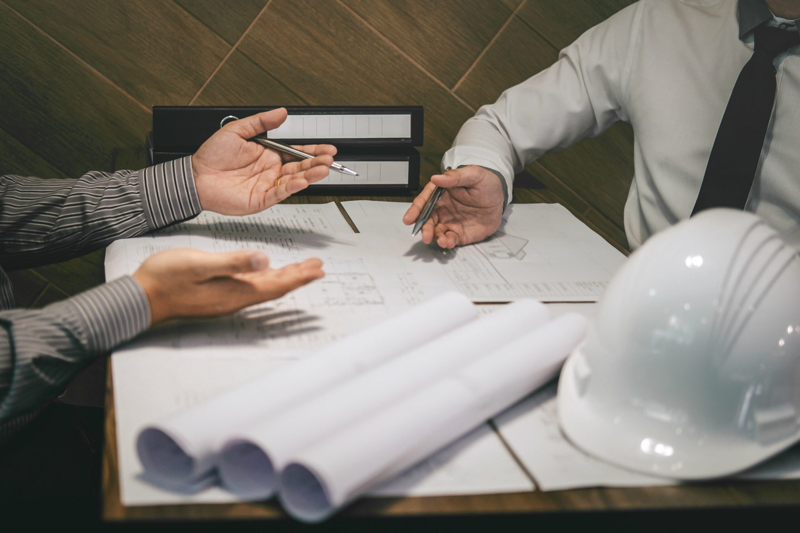 Two professionals discussing architectural blueprints and documents at a table with a white hard hat and rolled plans.