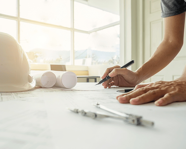 Person drawing on architectural blueprints at a table with a hardhat, rolled plans, and drafting compass nearby.