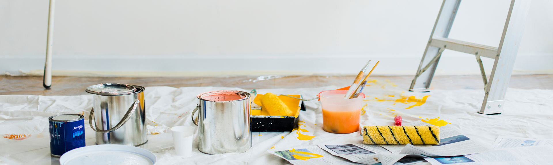Open paint cans, paint rollers, brushes in a plastic cup, and a metal ladder on a floor covered with a protective sheet during home painting.