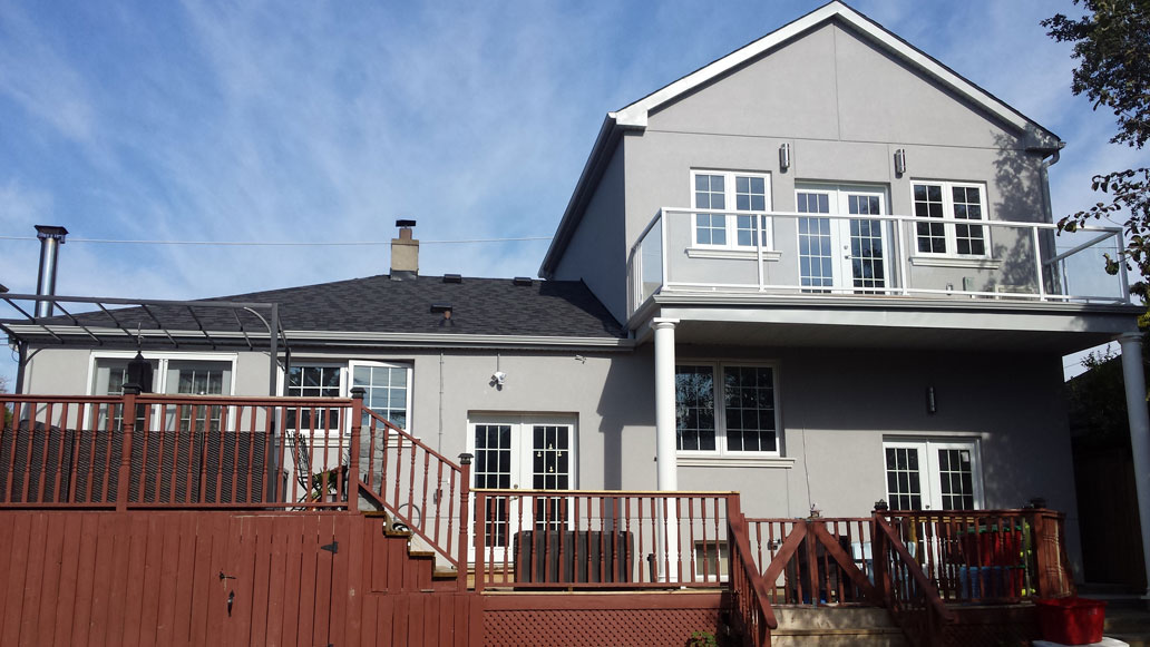 Two-story gray house with white trim, featuring multiple windows, a balcony with glass railing, and a wooden deck with stairs and railing in front.