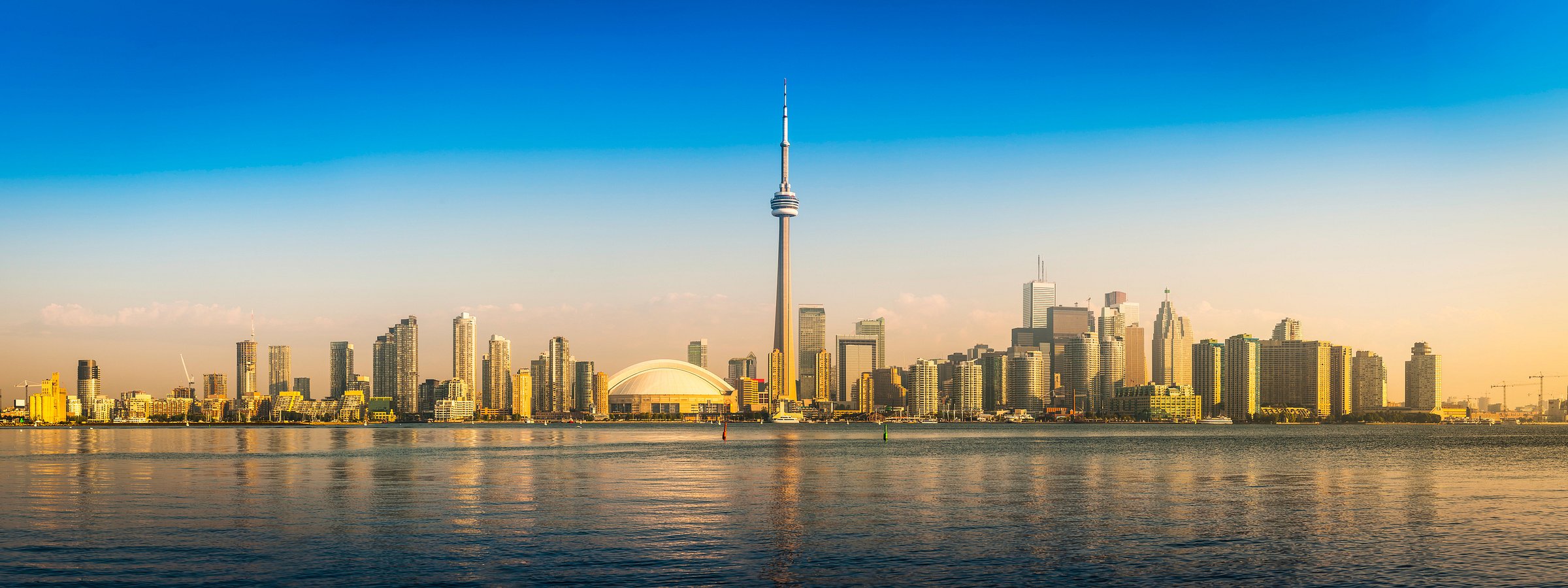 Toronto skyline with CN Tower and Rogers Centre dome at sunset reflecting on calm water.