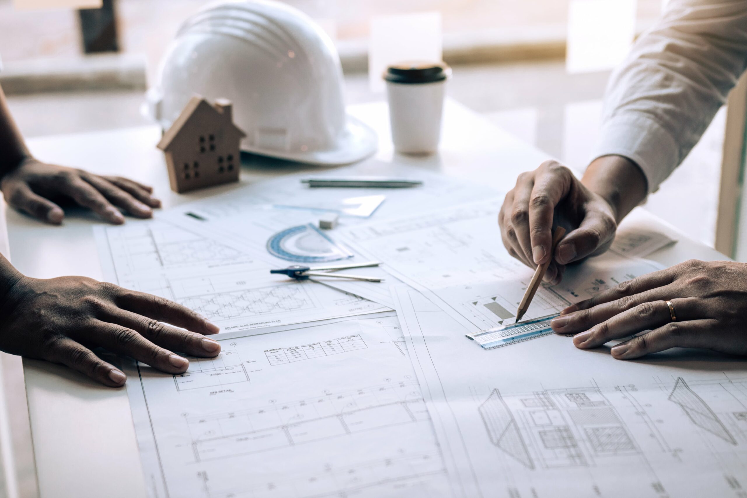 Hands of two people working on architectural blueprints with a ruler, compass, hard hat, and small house model on the table.