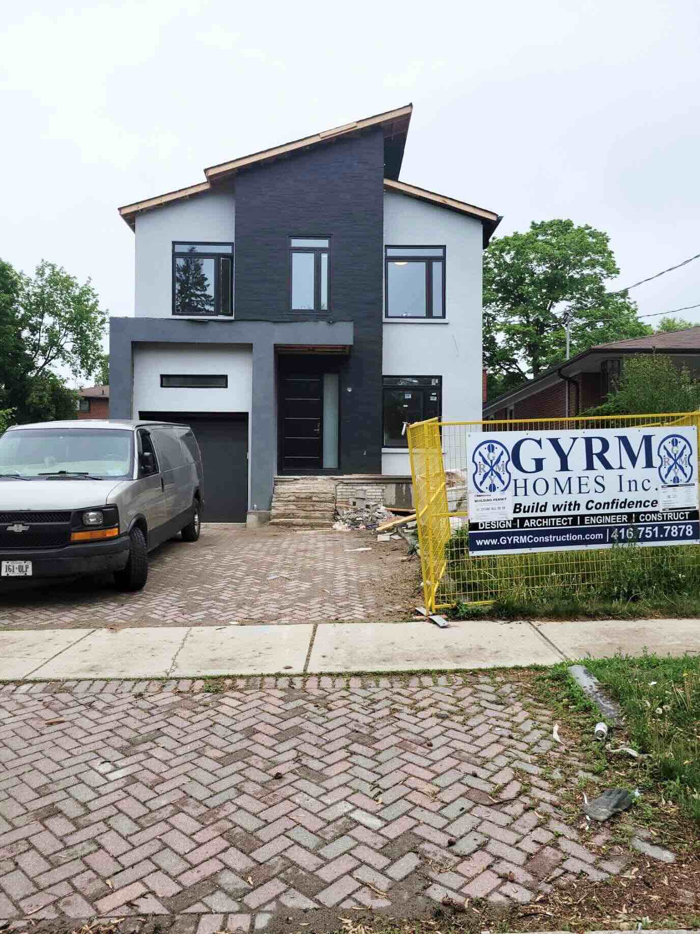 Modern two-story house with dark gray and white exterior under construction, a gray van parked in driveway, and a yellow fence with GYRM Homes Inc. construction sign.