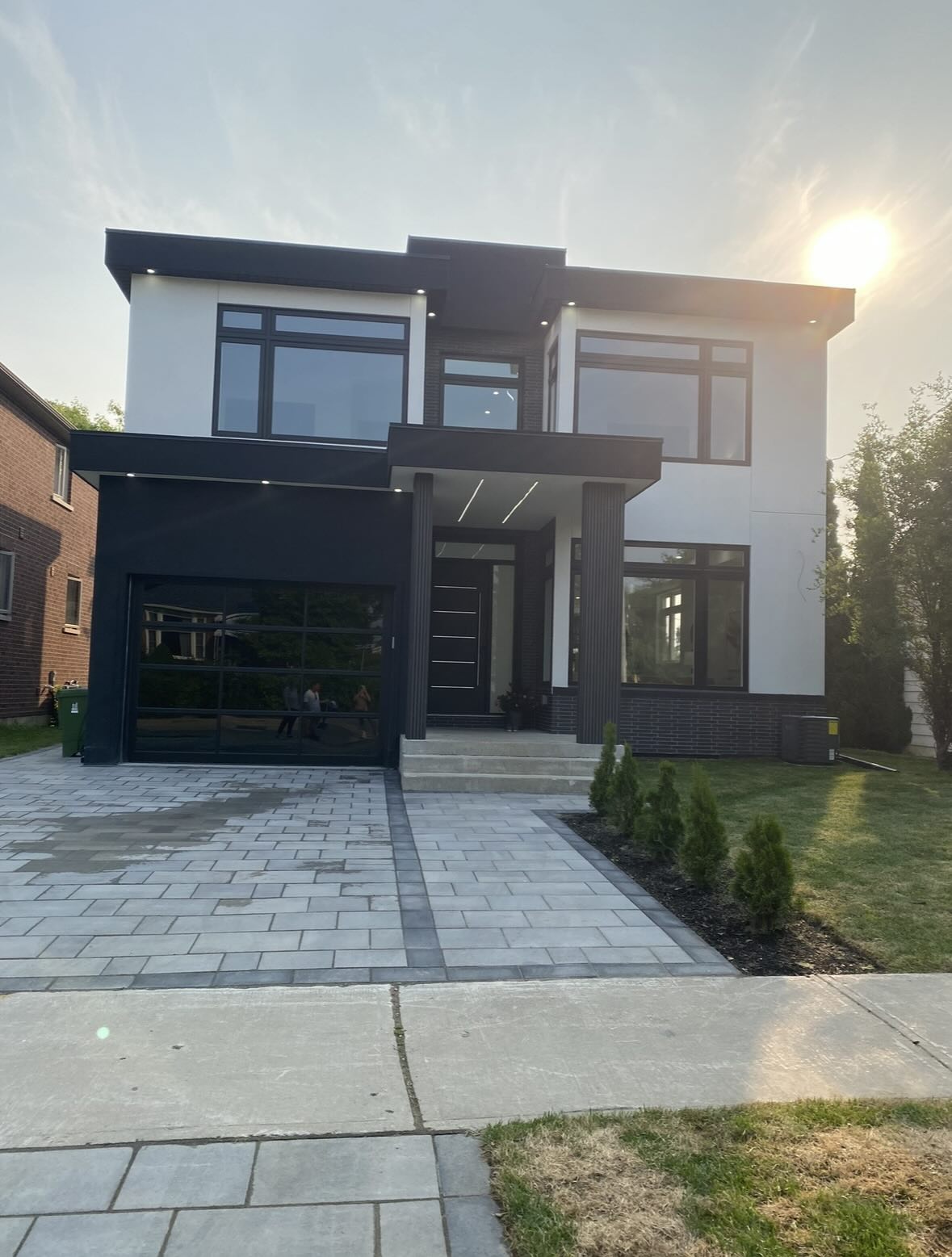 Modern two-story house with large windows, black and white exterior, a glass garage door, and a paved driveway with small shrubs on the side.