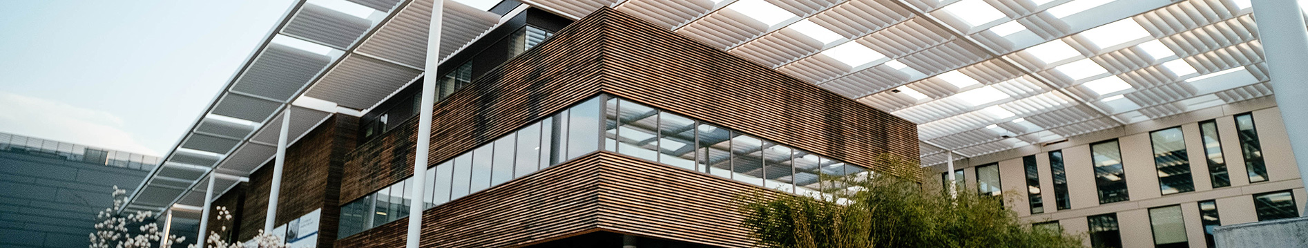 Modern building with wooden paneling and large windows under a white slatted canopy with greenery in front.