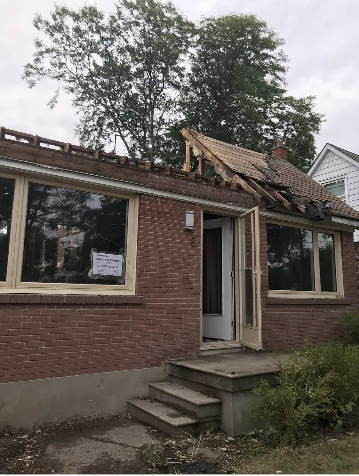 Brick house with partially removed roof and open door, showing renovation work and a building permit posted on a window.