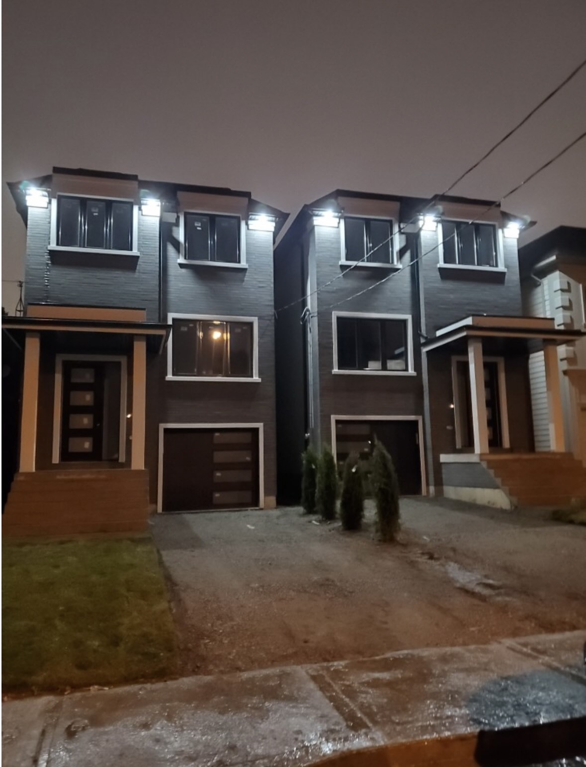 Night view of two modern semi-detached two-story houses with exterior lights on and small trees in front driveways.