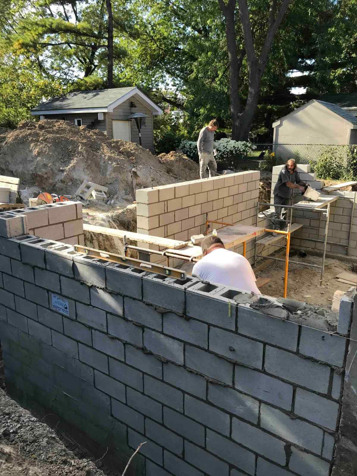 Three construction workers building walls with concrete blocks at an outdoor worksite surrounded by trees and small sheds.